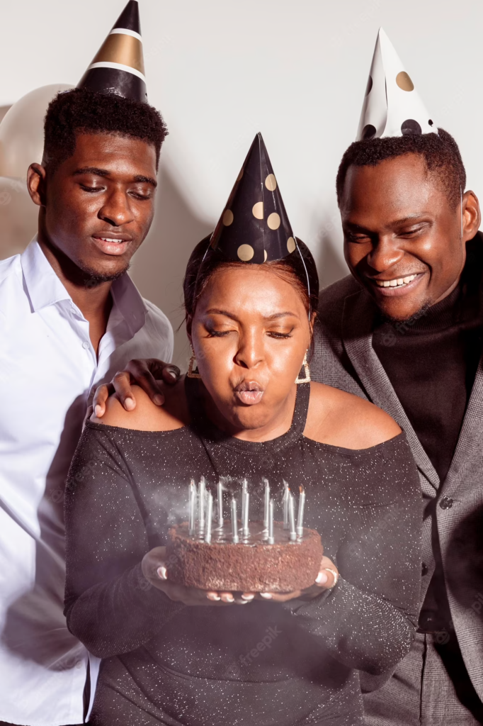 Black lady blowing a birthday cake, with her two male friends by her side.